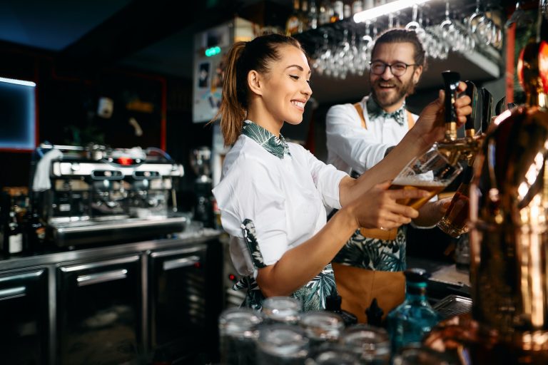 Female bartender pouring beer draft beer while working with colleague in a bar.